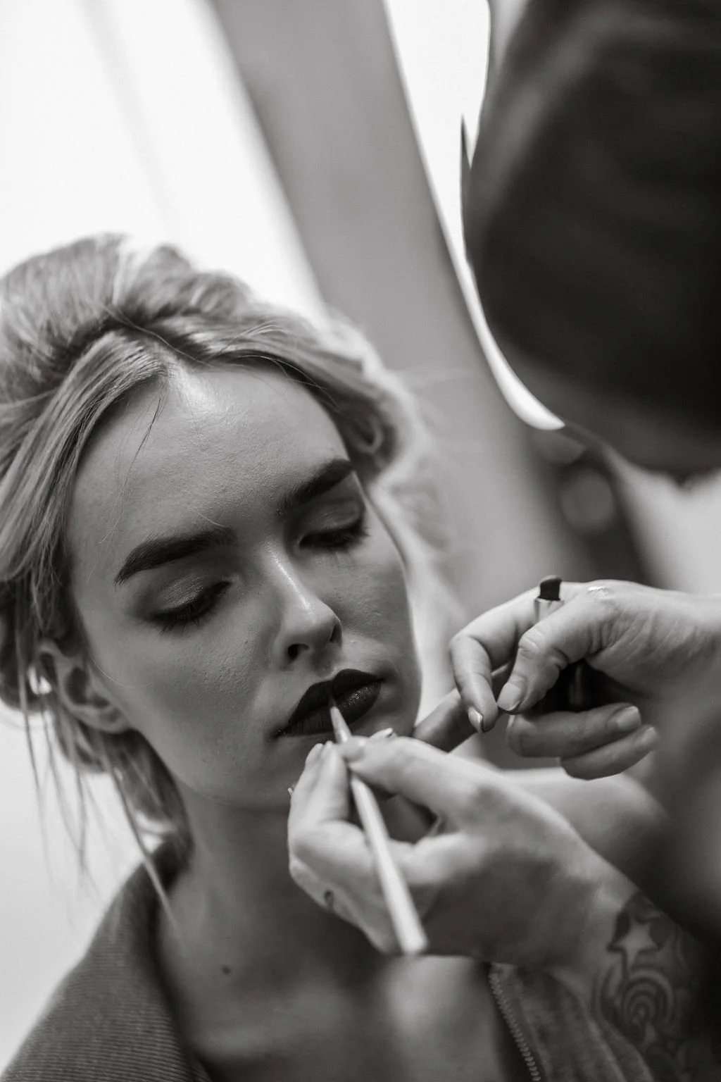 Black and white photo of a young woman with closed eyes getting her lips outlined with makeup by a makeup artist.
