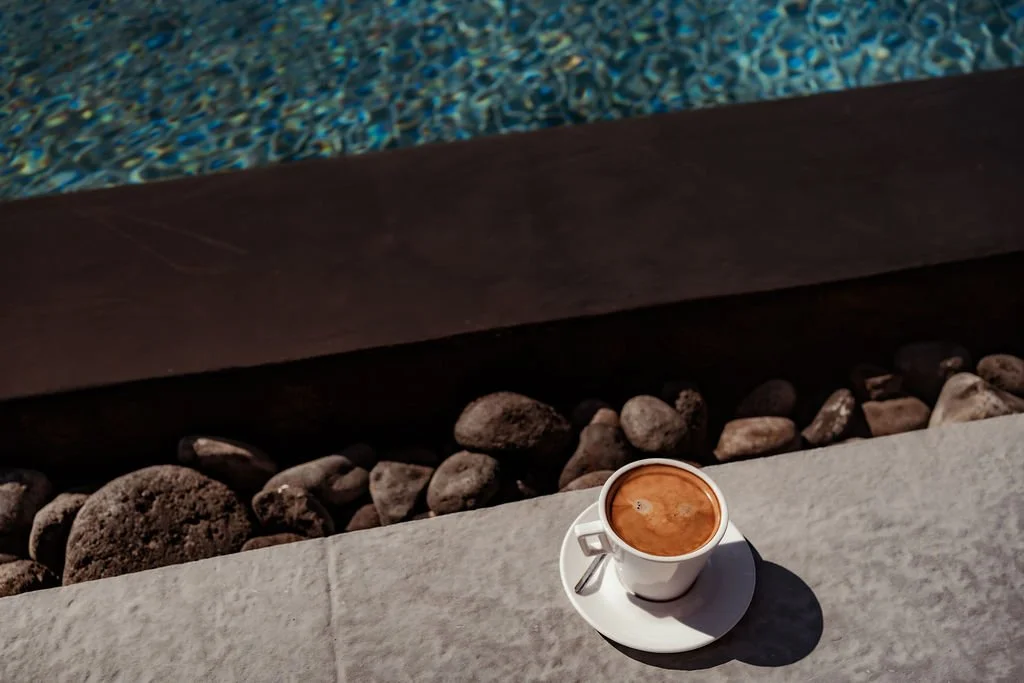 A cup of coffee on a saucer placed on a stone surface near a swimming pool, with rocks and the pool's edge visible in the background.