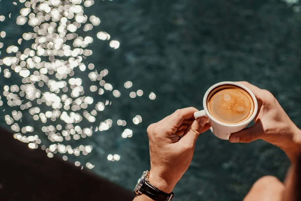 A person holds a white mug filled with coffee near a body of water with reflections of sunlight.