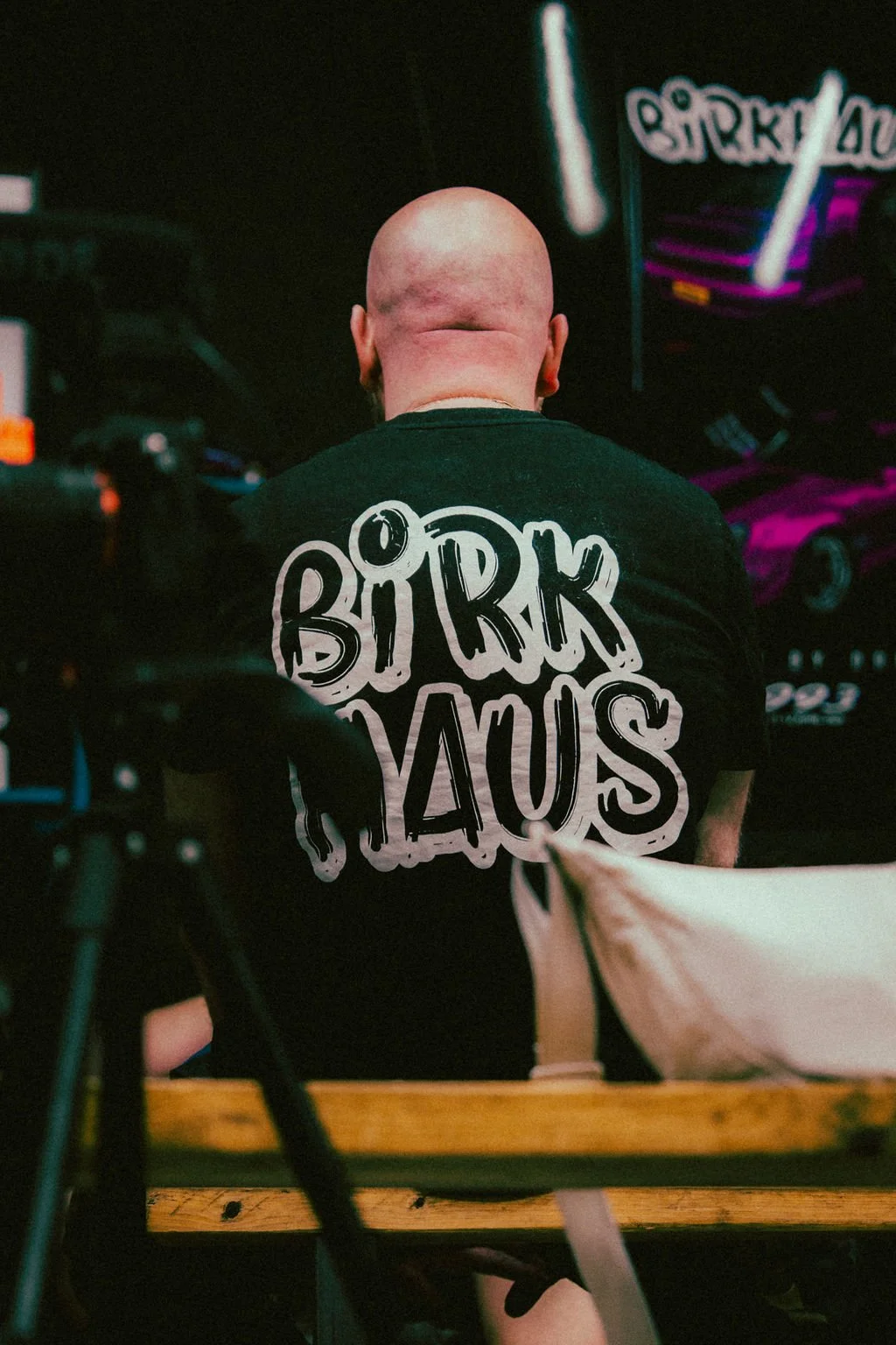 A person with a shaved head wearing a black t-shirt with white graffiti-style text that reads 'BIRK HAUS,' sitting with their back to the camera. The scene appears to be indoors with dark and colorful lighting, possibly in a studio or a nightclub.