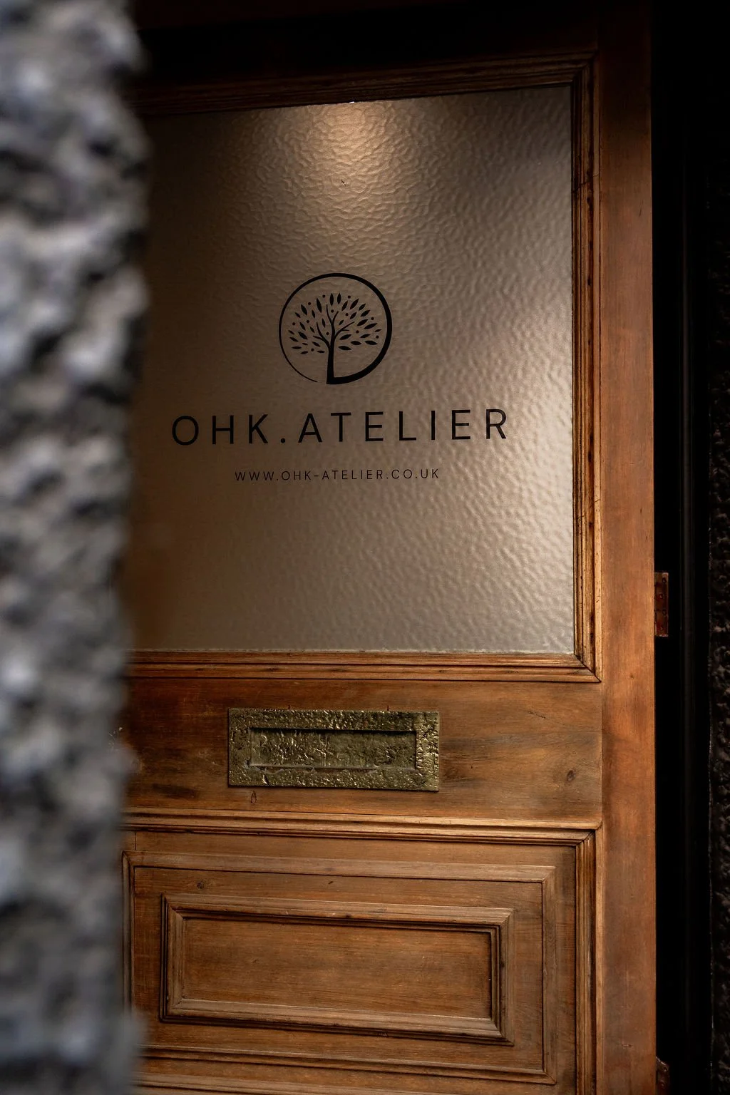 Wooden door with a frosted glass window displaying the logo and website of OHK.ATELIER, featuring a tree icon and text.
