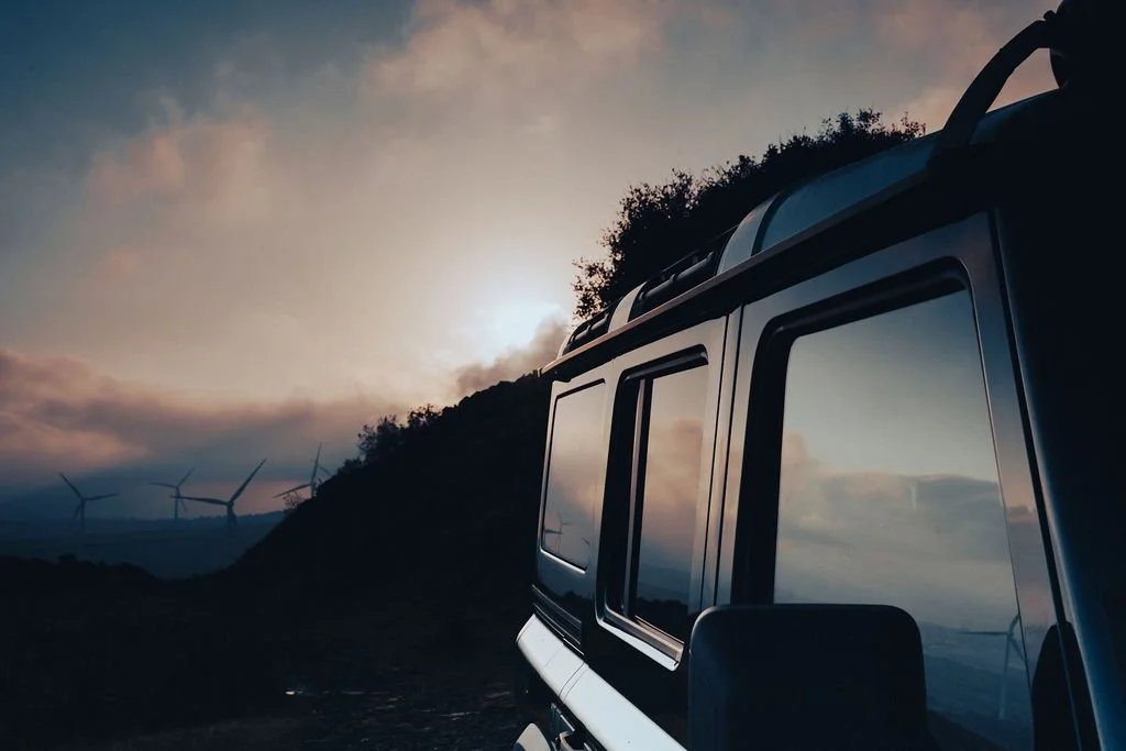 A reflection of cloudy sky on the side window of a dark-colored vehicle, with wind turbines on a hill in the background at sunset.