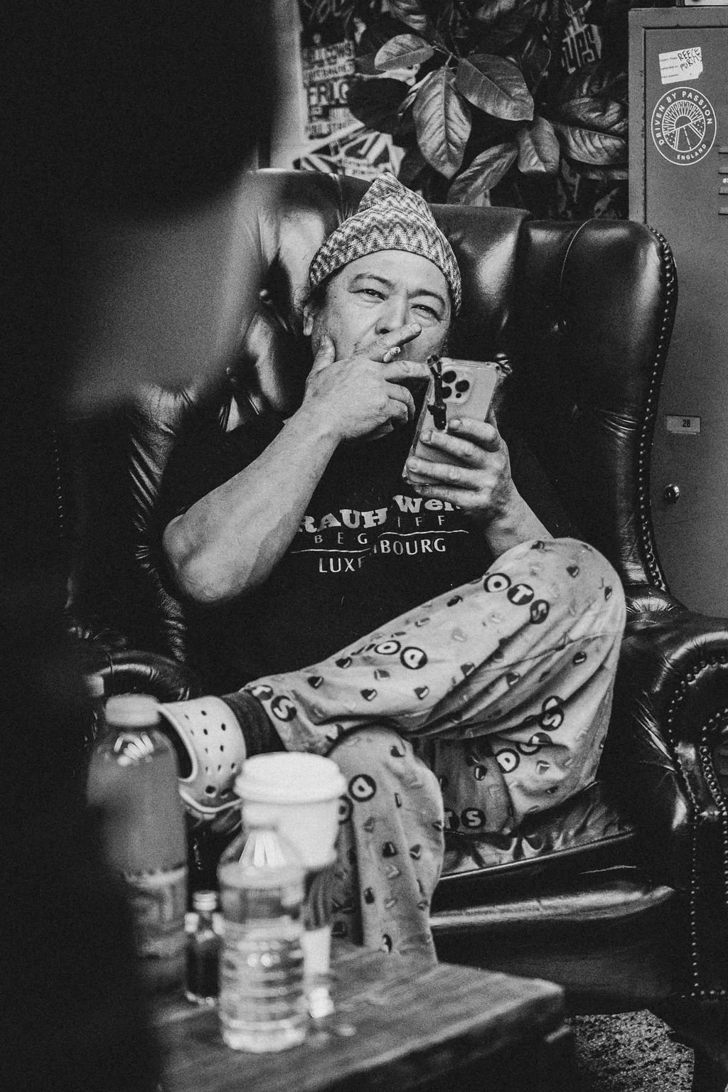 A man sitting in a leather armchair, wearing a beanie and patterned pajama pants, holding a smartphone and making a gesture near his face. There are bottles and a coffee cup on a table in front of him, and a large plant and lockers in the background.