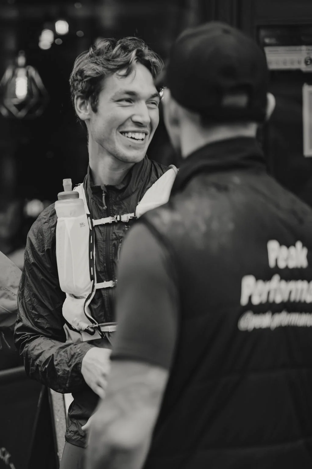 Two young men talking, one smiling, in athletic gear, at an event, with one wearing a jacket that has words "Peak Performance" on the back.