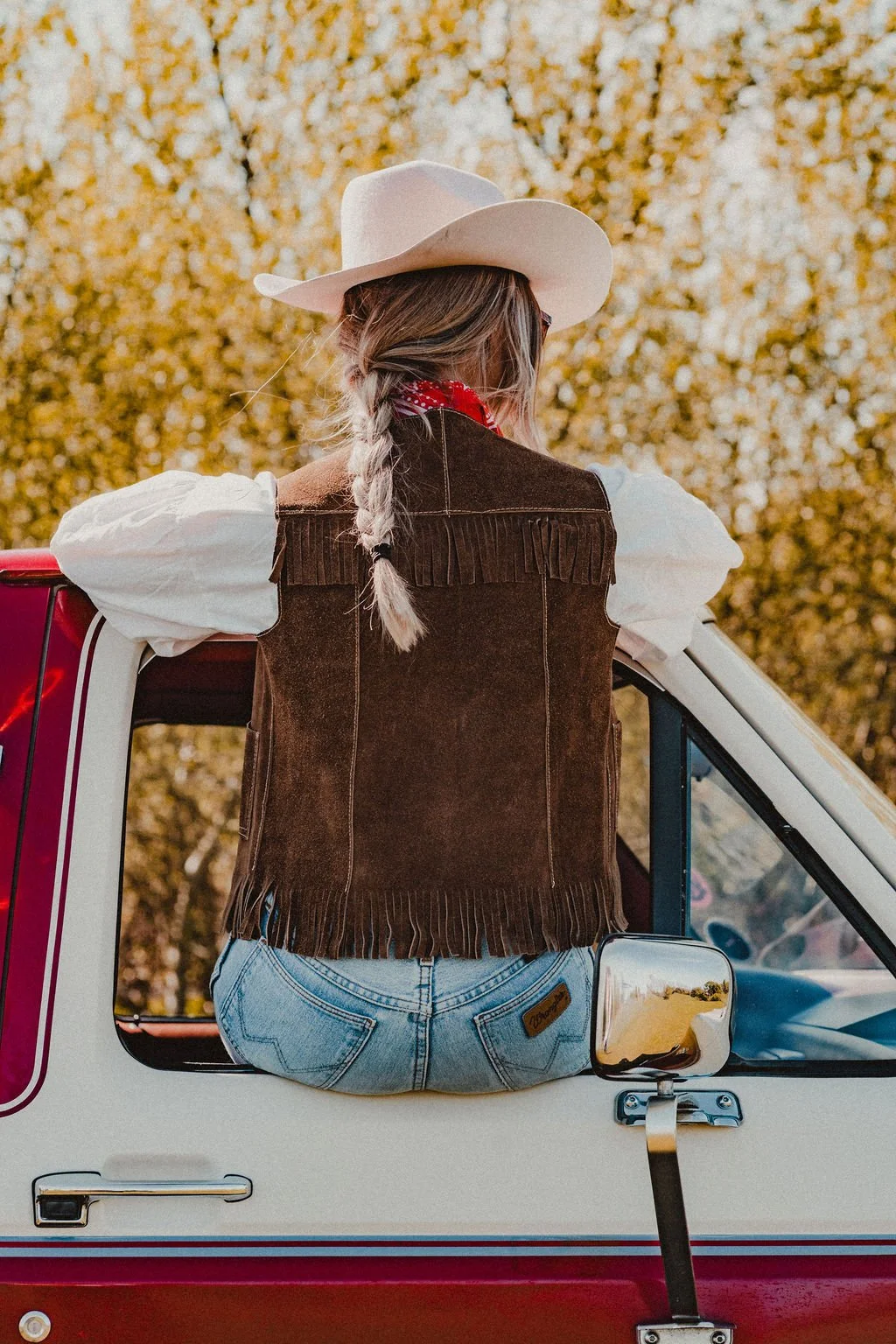 Woman wearing a cowboy hat, brown fringed vest, white blouse, and jeans, sitting on the open door of a vintage red and white pickup truck, with fall foliage in the background.