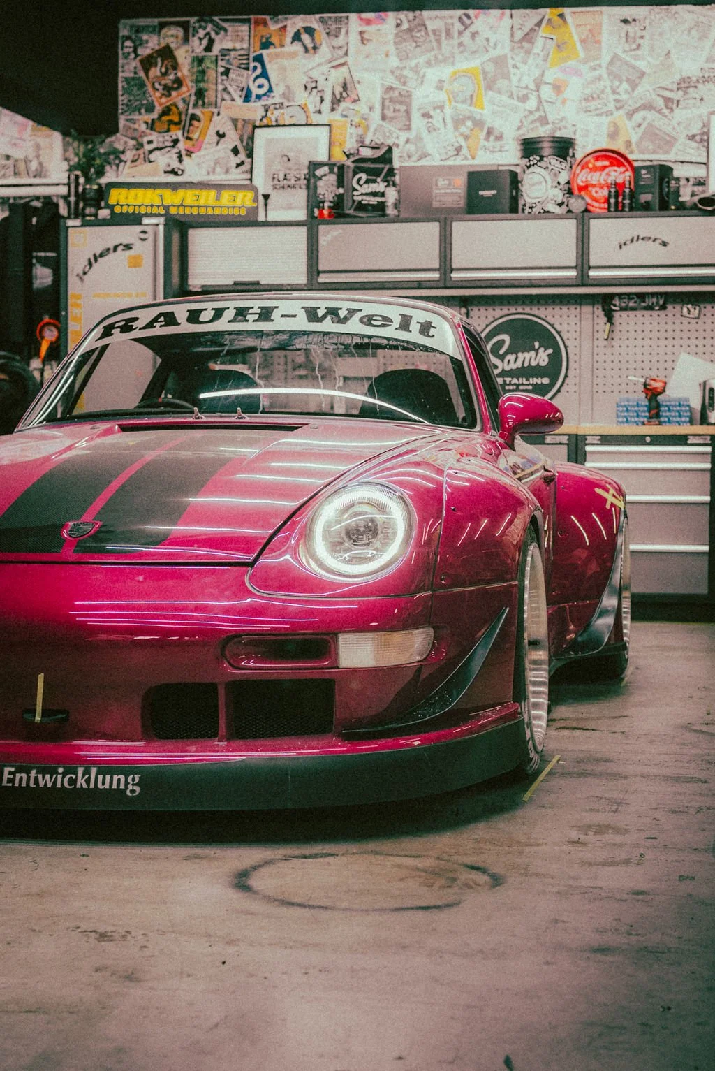 A pink Porsche RWB with black accents inside a garage, surrounded by tools, signs, and automotive memorabilia.