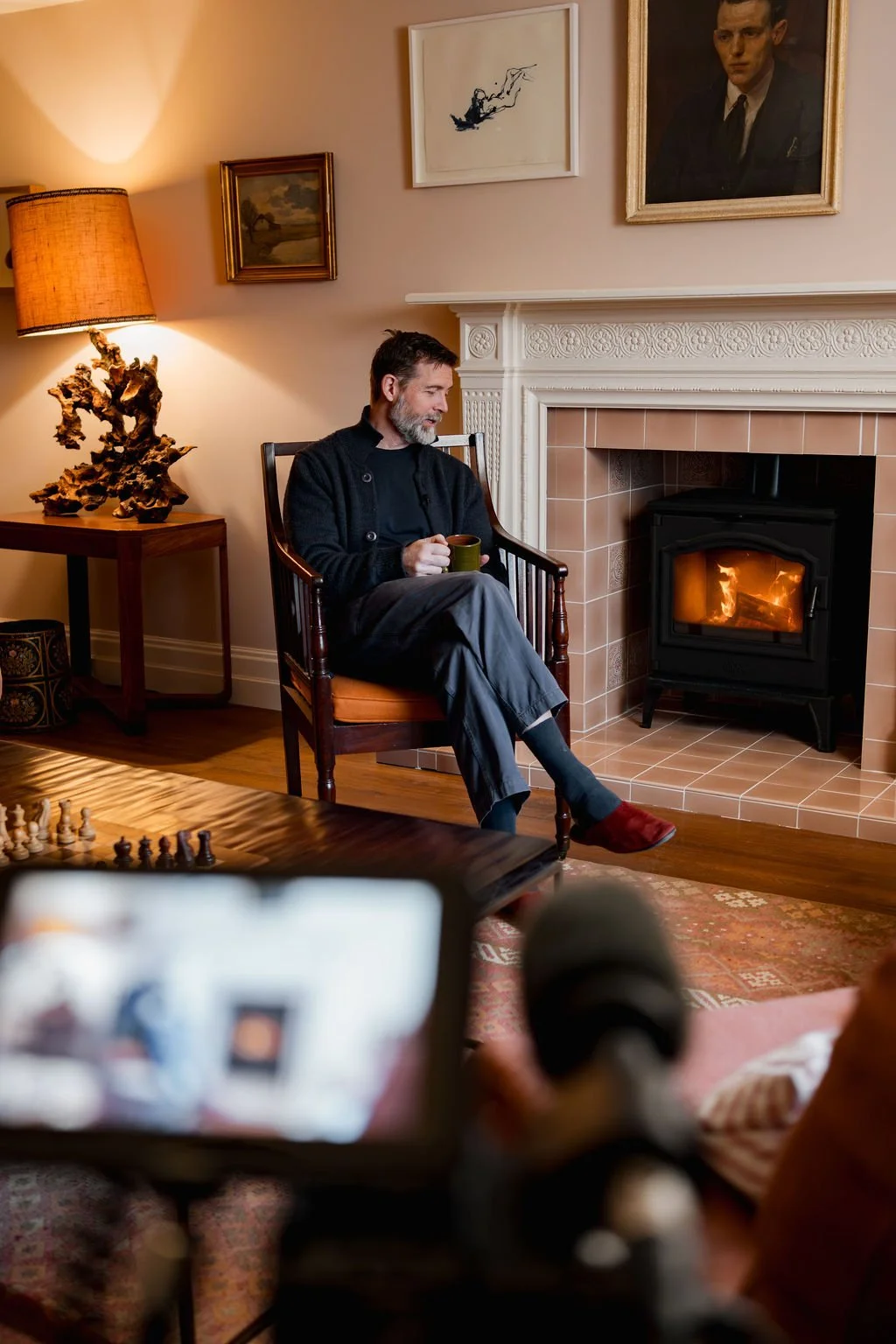 A man sitting in a wooden chair with a cushion, holding a mug and relaxing by a fireplace in a cozy living room, decorated with framed paintings and warm lighting.