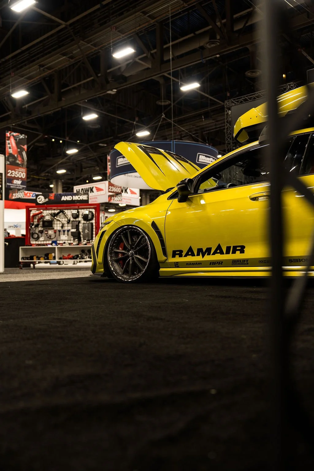 Yellow race car with hood open at an indoor automotive event, with various car parts displayed in the background.