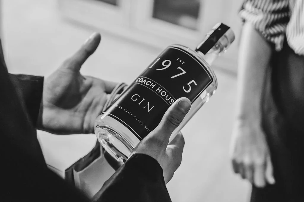 A person's hands holding a large gin bottle labeled '1975 Coach House Gin'. The background is blurred.