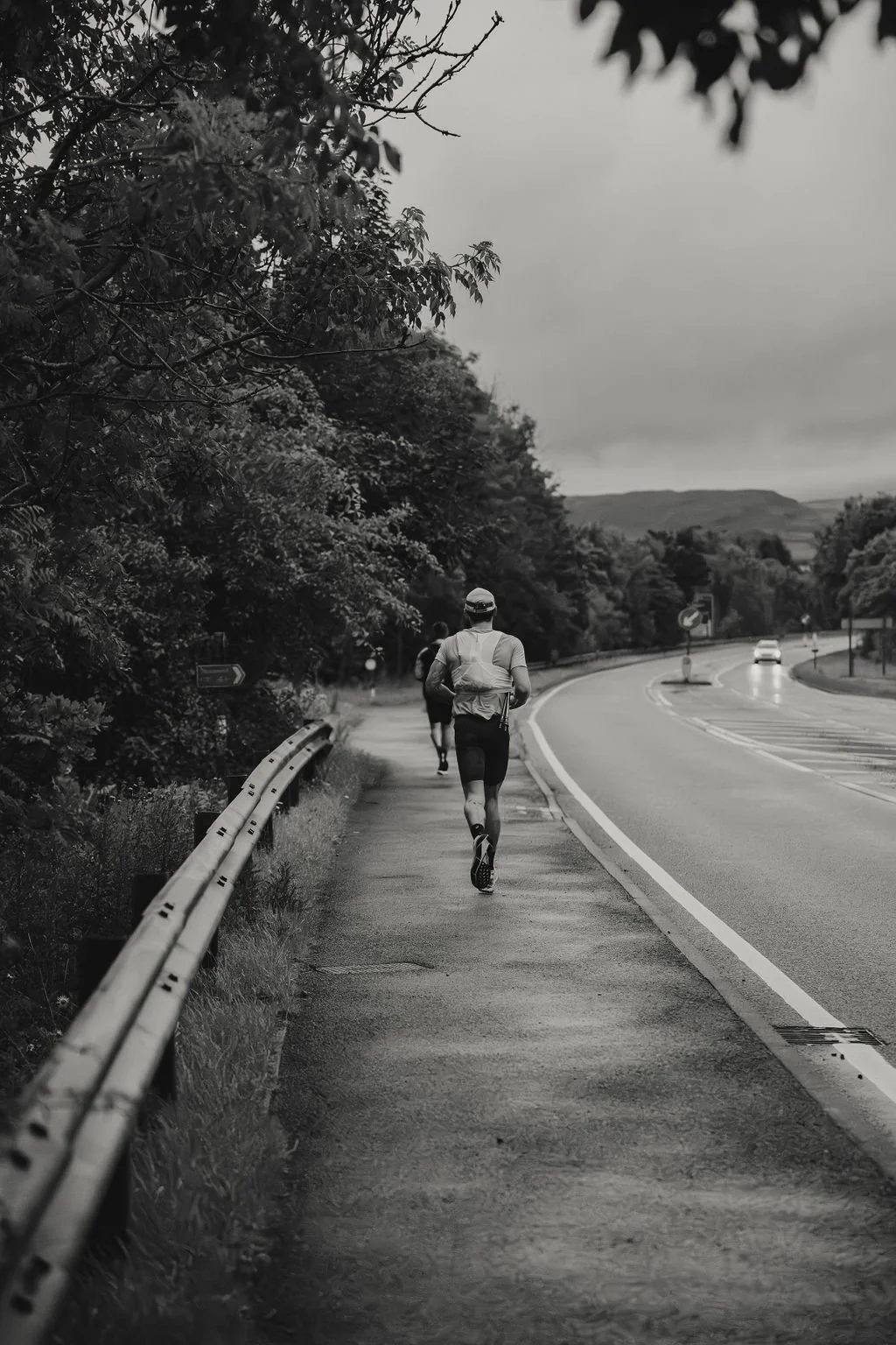 A man running along a roadside path on a cloudy day, with another runner in the background and cars on the road to the right, surrounded by trees and hills.