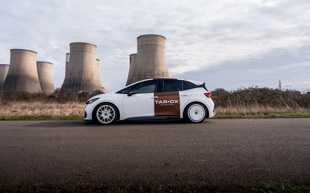 A white hatchback car with custom wheels and a Taro X advertising wrap parked on the side of a road near a row of cooling towers at a power plant, under a cloudy sky.