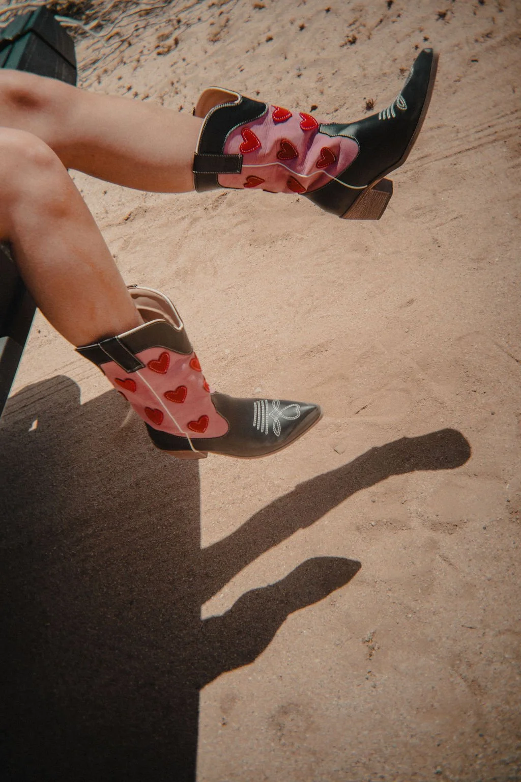 Close-up of a person's legs wearing colorful cowboy boots with red hearts, sitting on sandy ground, with their shadow visible on the sand.
