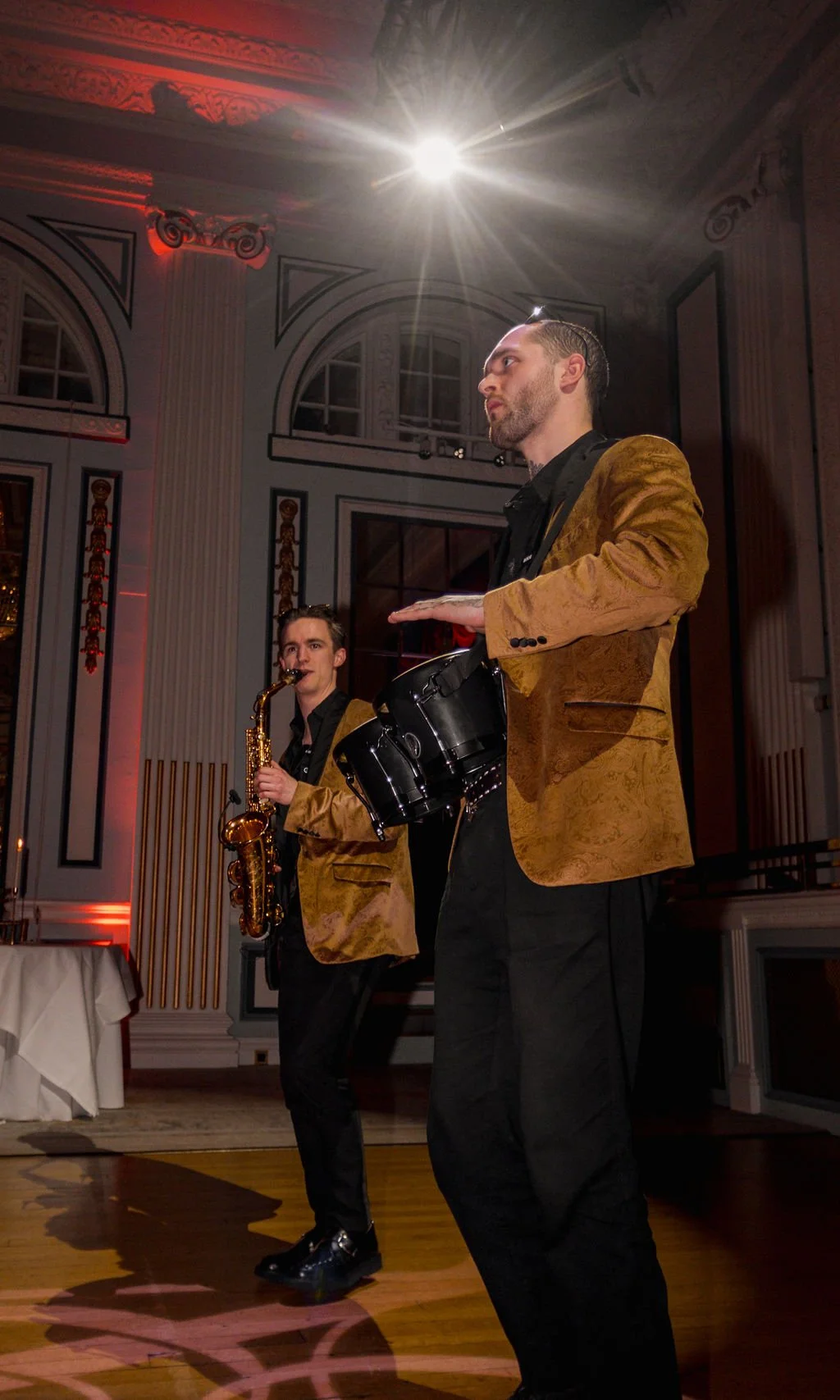 Two musicians performing at an elegant event, one playing a saxophone and the other playing a drum, in a decorated hall with ornate columns and large windows.