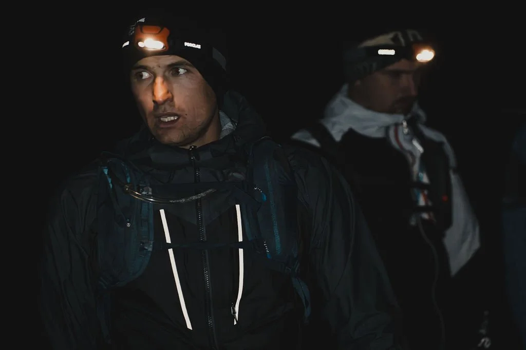 Two men wearing headlamps and outdoor gear at night, possibly preparing for or during an adventure or expedition.