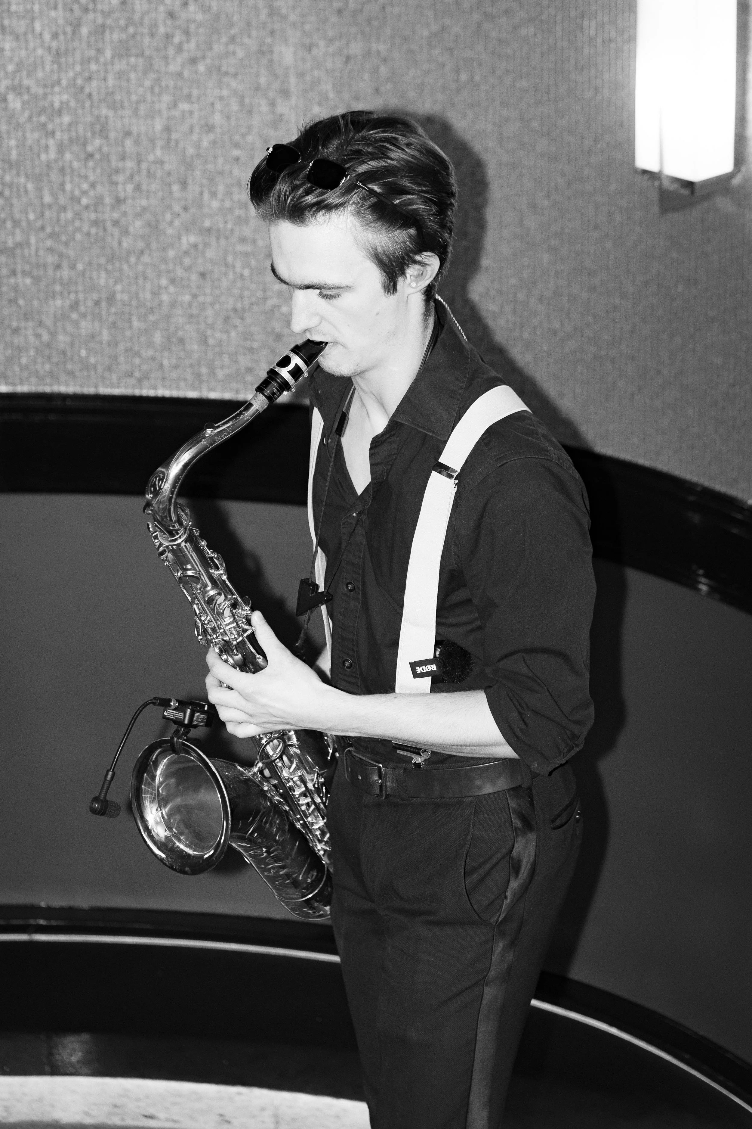 A young man playing the saxophone indoors, dressed in black with glasses pushed up on his head.