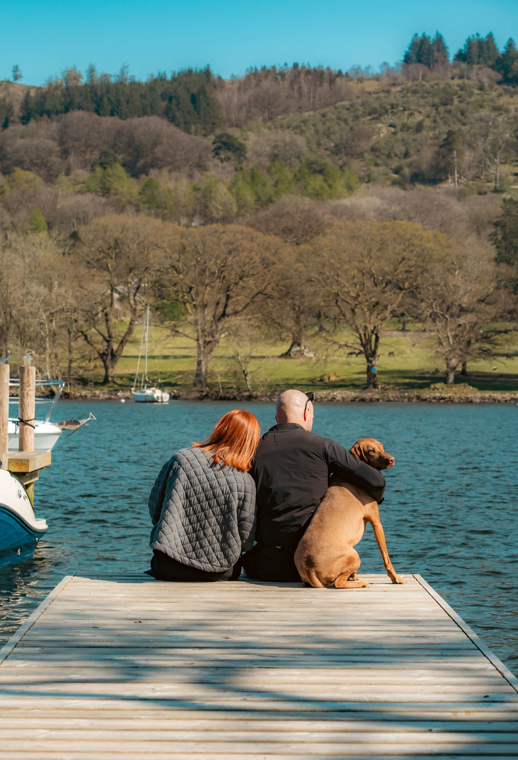 A man and a woman sitting on a wooden dock by a lake with a brown dog. They are facing away from the camera, looking at the water and trees in the distance.