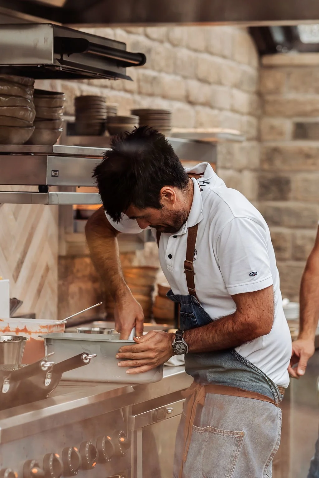 A man wearing a white polo shirt and an apron is cooking in a restaurant kitchen, leaning over a stove with pots and pans around him.