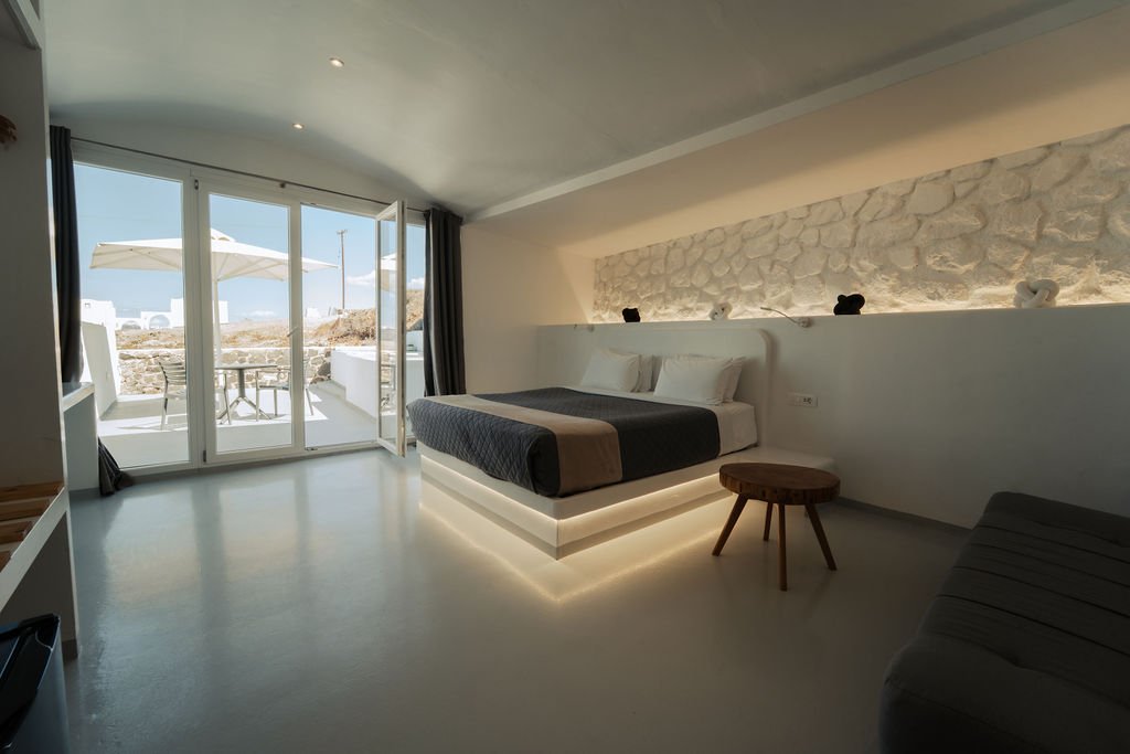 Modern bedroom with a large glass door leading to a balcony with outdoor furniture and umbrella, white walls with stone accent wall, bed with white headboard and dark bedding, small wooden stool, and gray seating to the right.