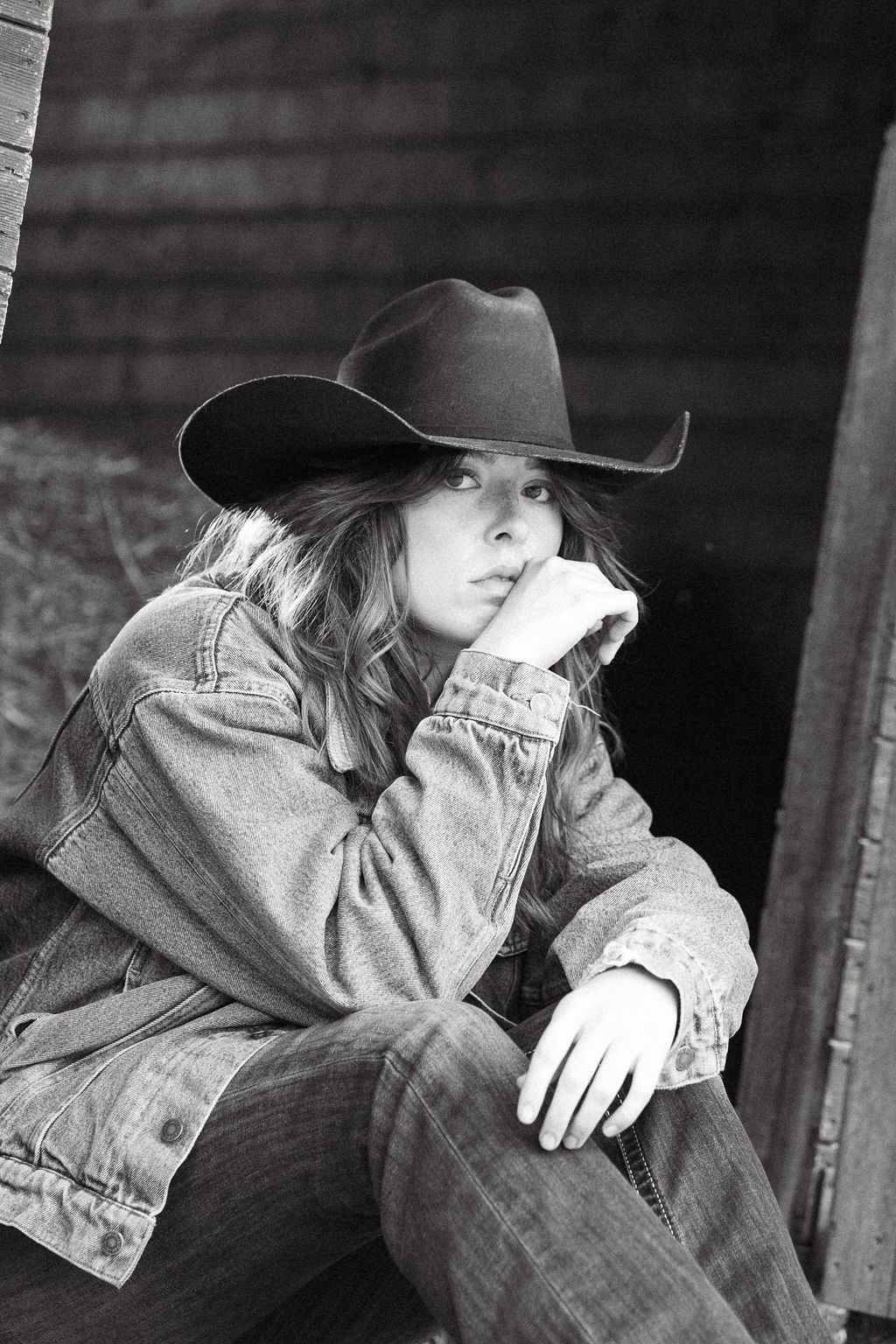 A young woman wearing a cowboy hat and denim jacket sitting outdoors near a wooden structure.