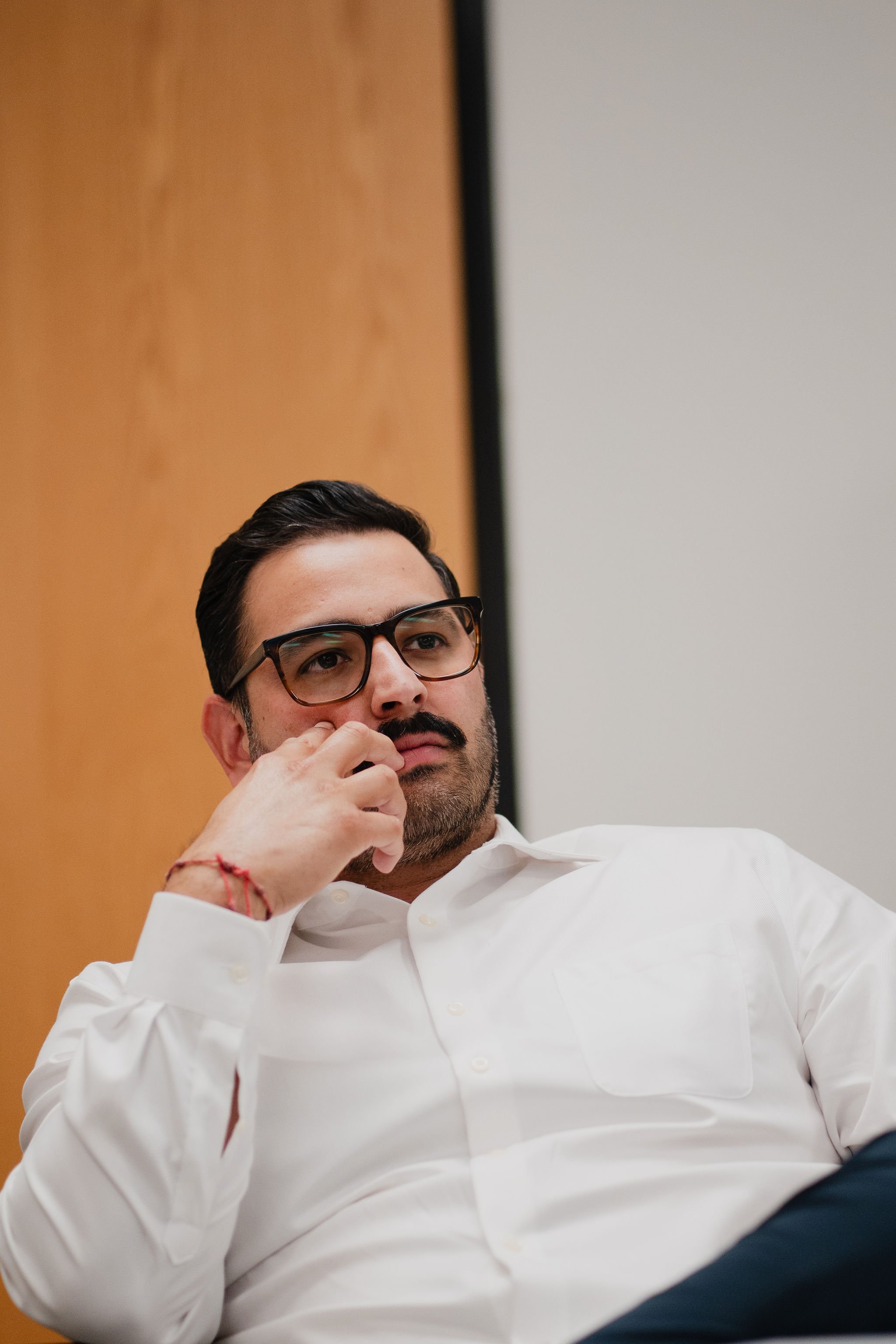 A man with glasses and a beard wearing a white shirt, sitting thoughtfully with his hand near his face.