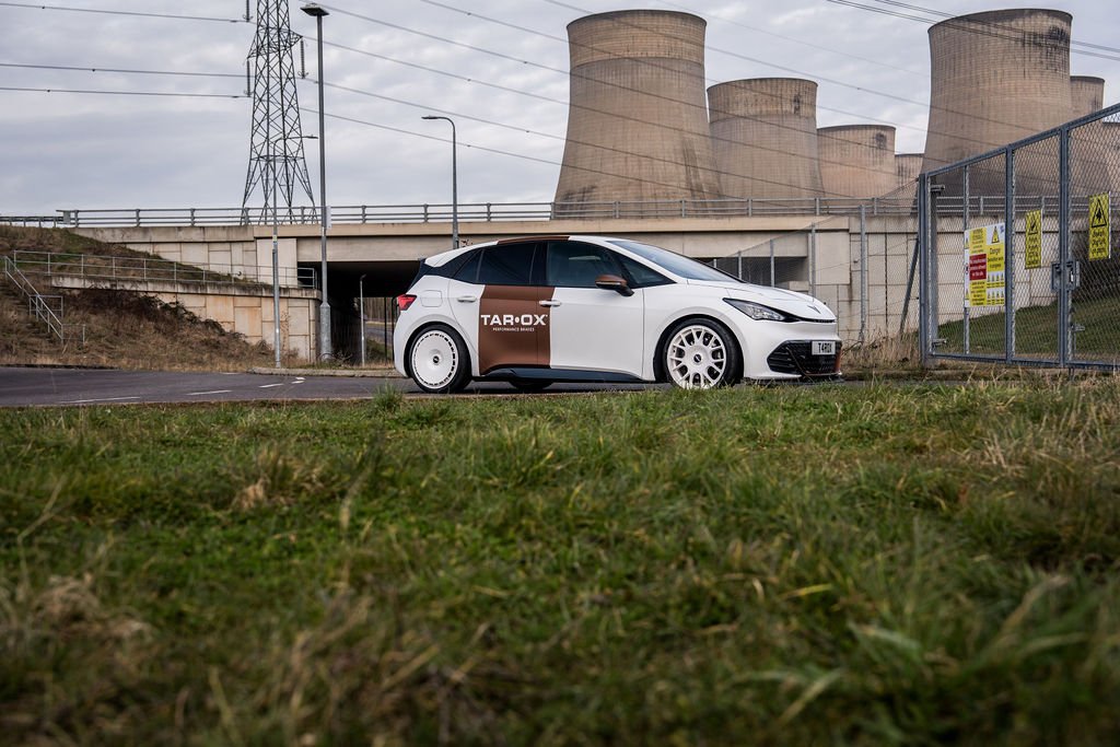 A white hatchback car with black and brown markings parked on a grassy area near a gated electrical substation with cooling towers in the background.