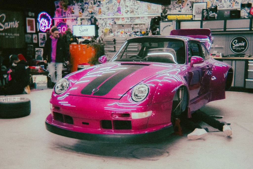 A pink Porsche RWB with black stripes in a garage, with a person working underneath it and another person standing nearby, surrounded by tools and accessories.