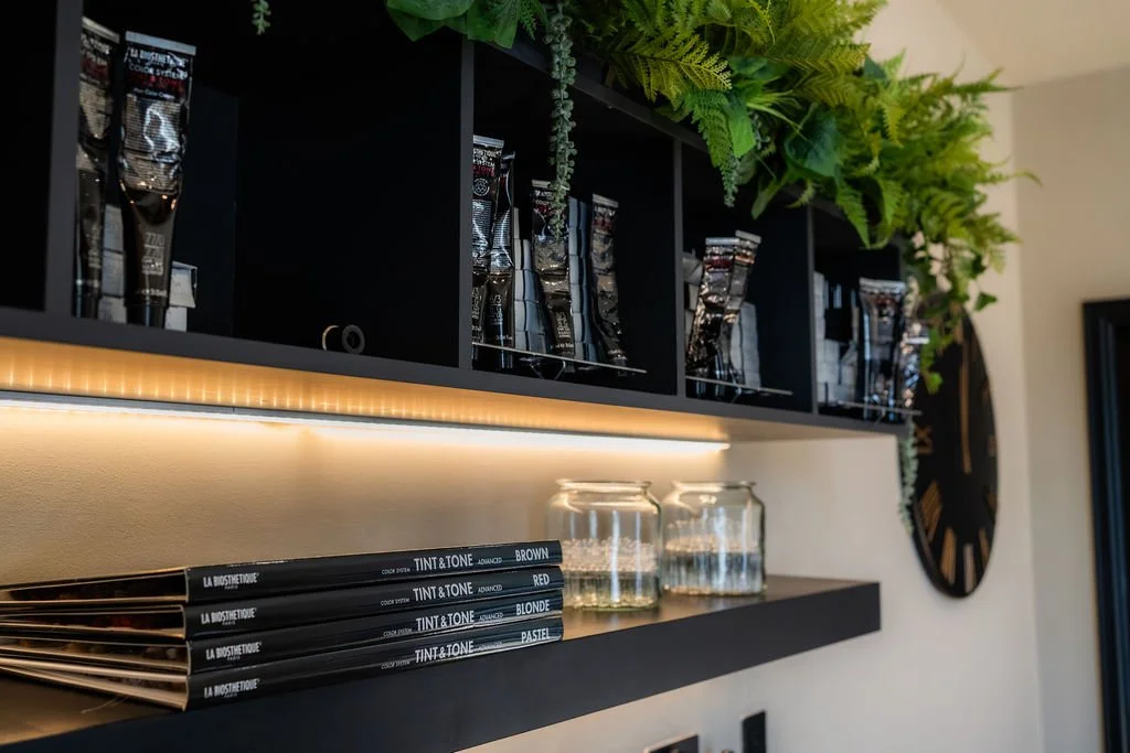 Black shelf with hair dye color swatch, glass jars, and manicure tools. Green plants above the shelf and a large clock on the wall.