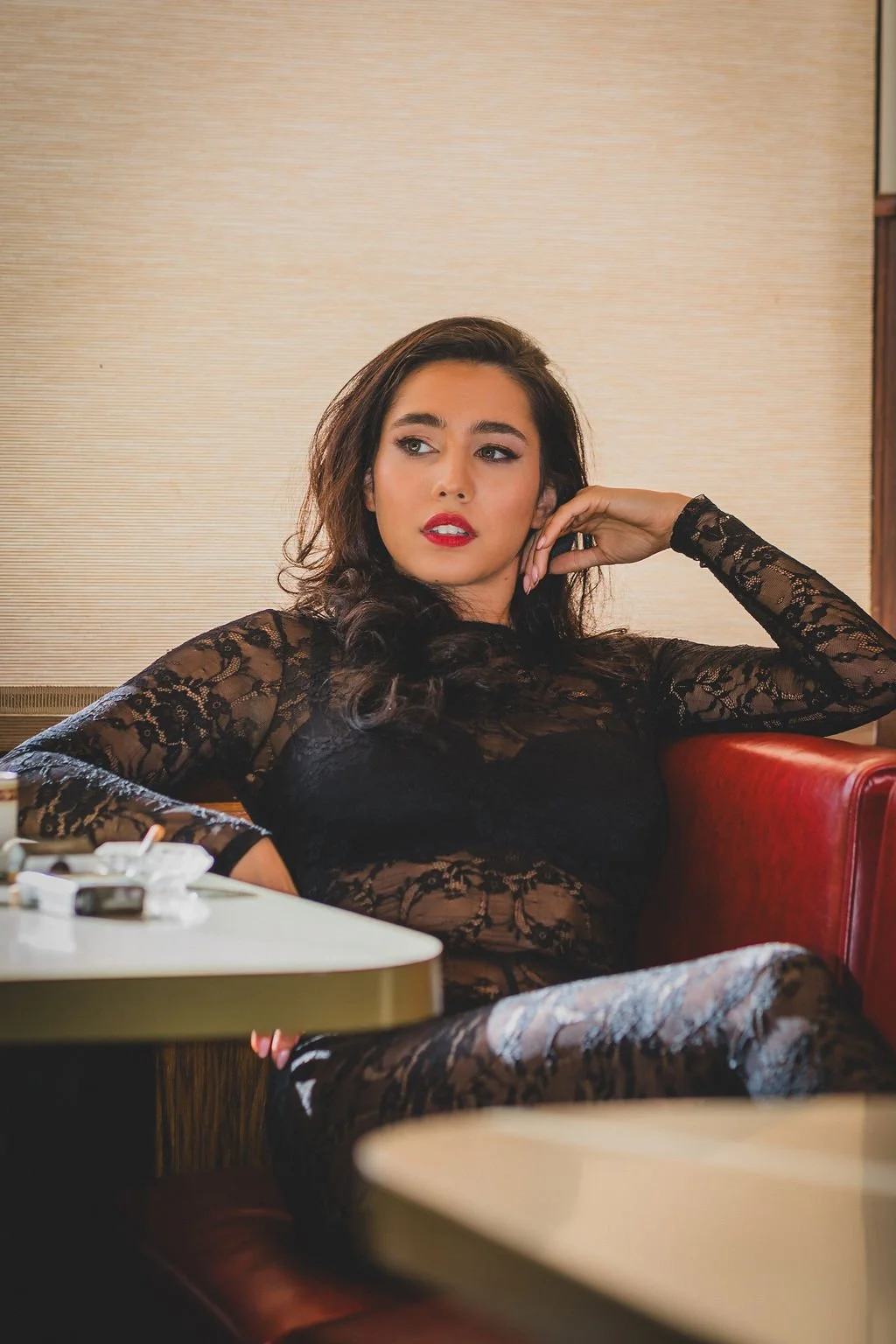 A young woman with dark wavy hair and red lipstick wearing a black lace lingerie, sitting in a restaurant booth with a serious expression, with cigarettes and a lighter on the table.