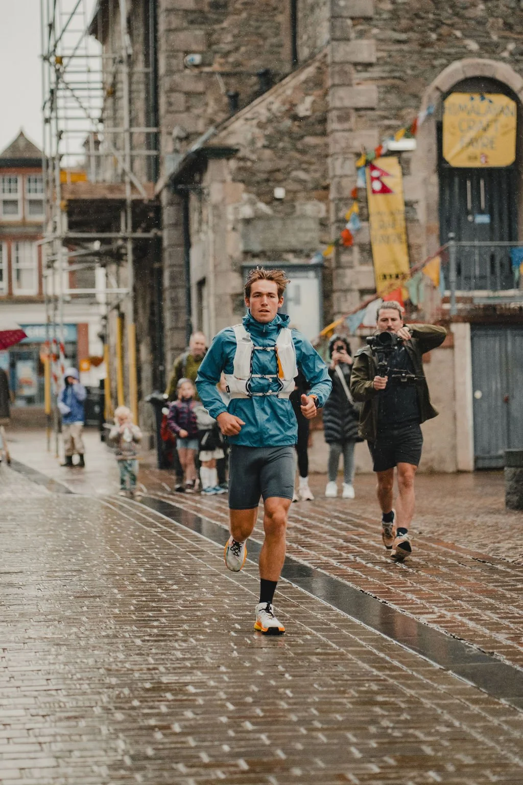 A man running on a wet cobblestone street in a city, wearing athletic clothing and a backpack, with several people and a cameraman in the background.