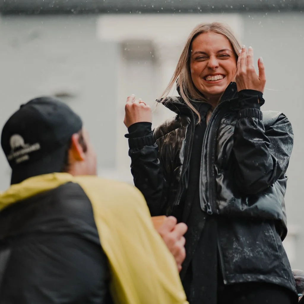 A woman with long blonde hair wearing a black jacket is smiling and touching her face, while a man wearing a black cap and black jacket with yellow accents is kneeling and looking at her.