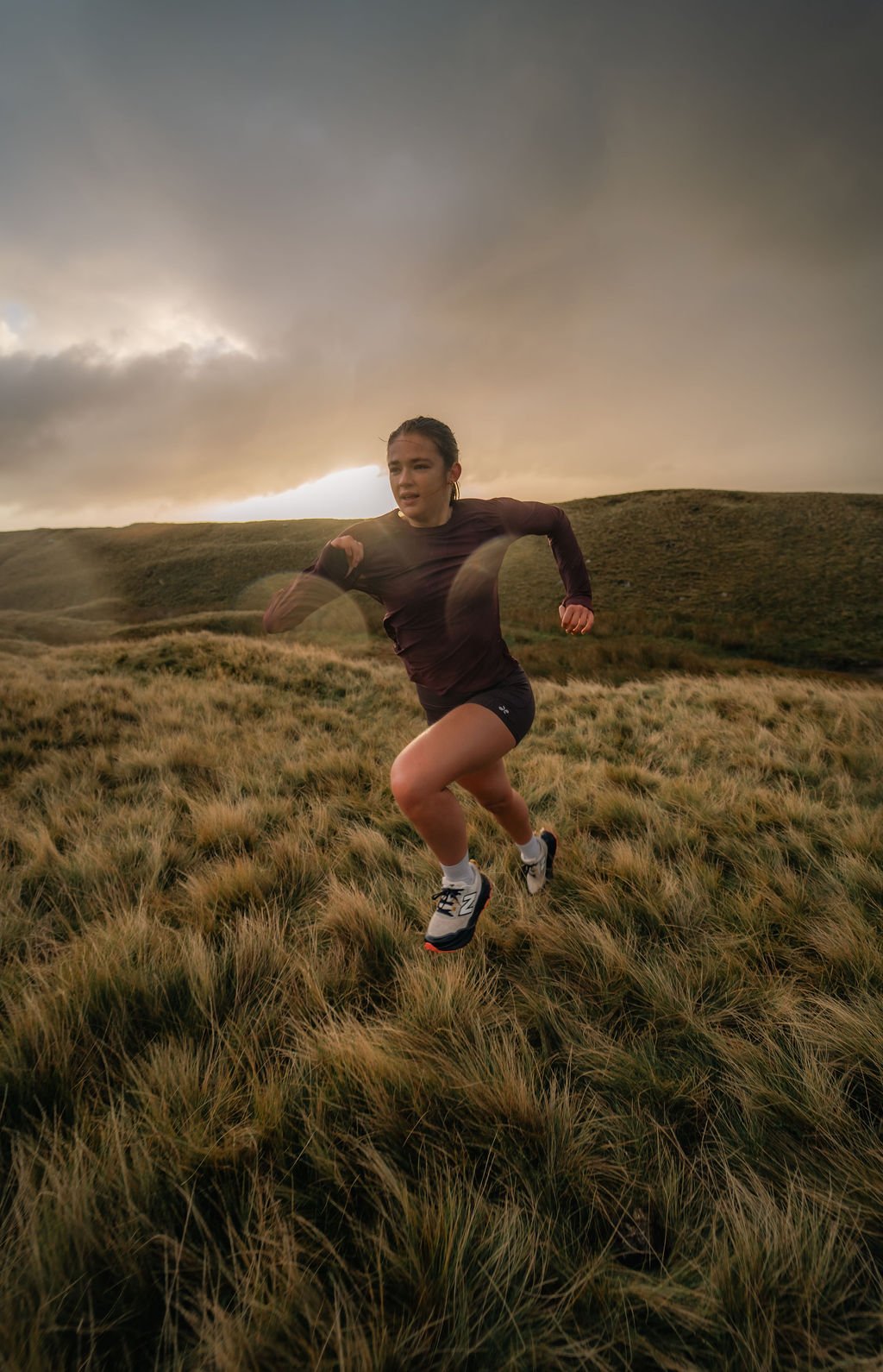 A woman running outdoors on grassy terrain with rolling hills and a dramatic cloudy sky in the background during sunset.