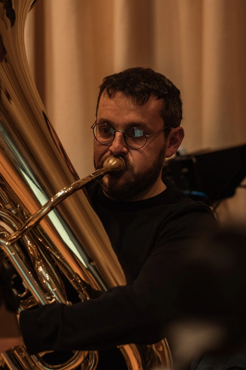 A man with glasses playing a brass euphonium or tuba, focused on the instrument with a curtain and piano in the background.