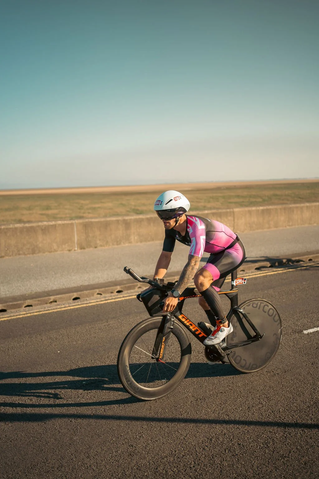 A male cyclist wearing a white helmet, sunglasses, and a pink and black cycling suit rides a black bike with disc wheels on a paved road through a flat landscape during the daytime.