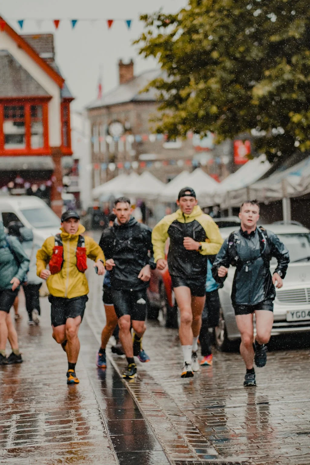 Group of runners participating in a race on a rainy day in a city street, with tents and buildings in the background.