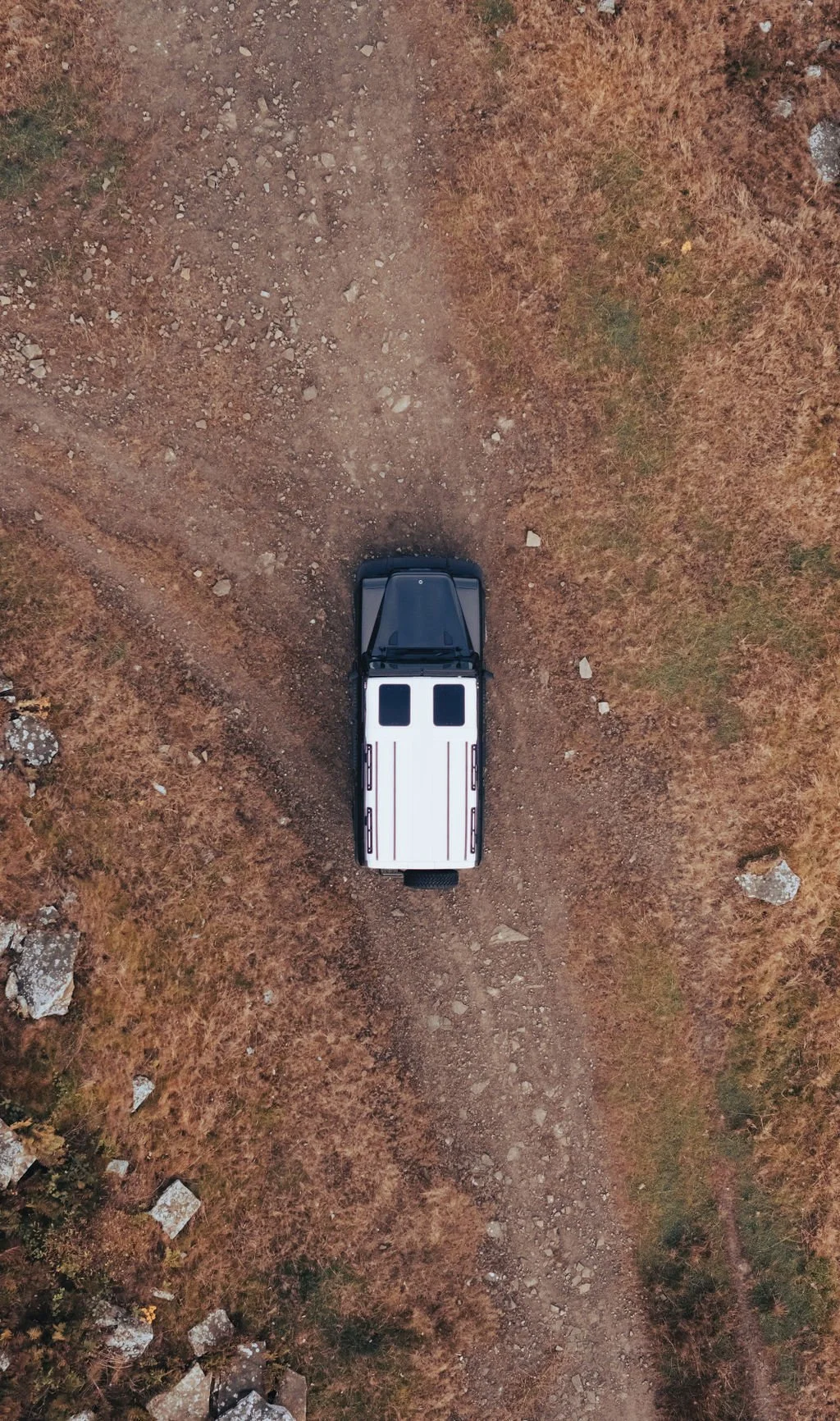 Overhead view of a black and white pickup truck on a dirt trail through a dry, grassy area with rocks scattered around.