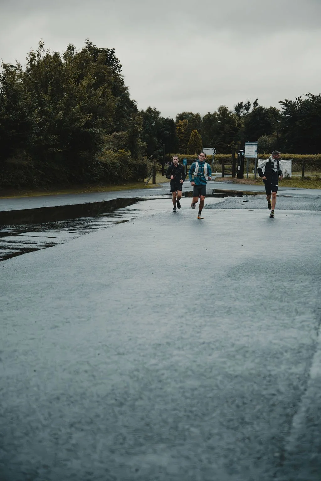 Three men running on a wet road in a park on a cloudy day, with trees and park signs in the background.
