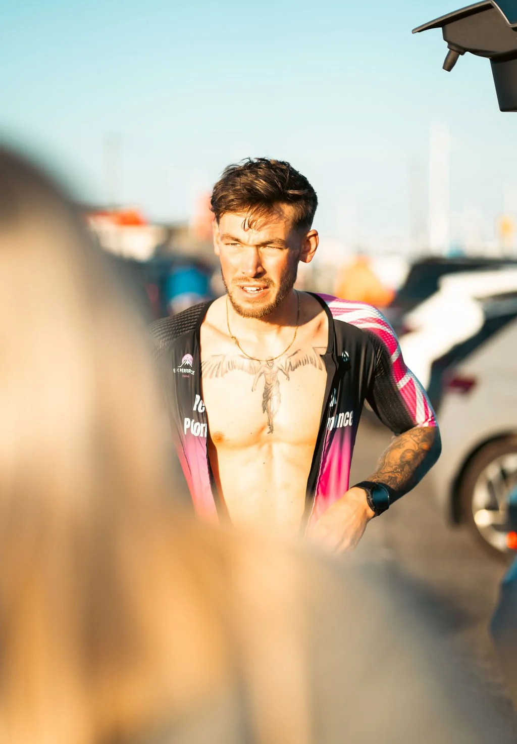 A young man with tattoos, wearing a black and pink sports jacket, is talking outdoors with parked cars and a clear blue sky in the background.