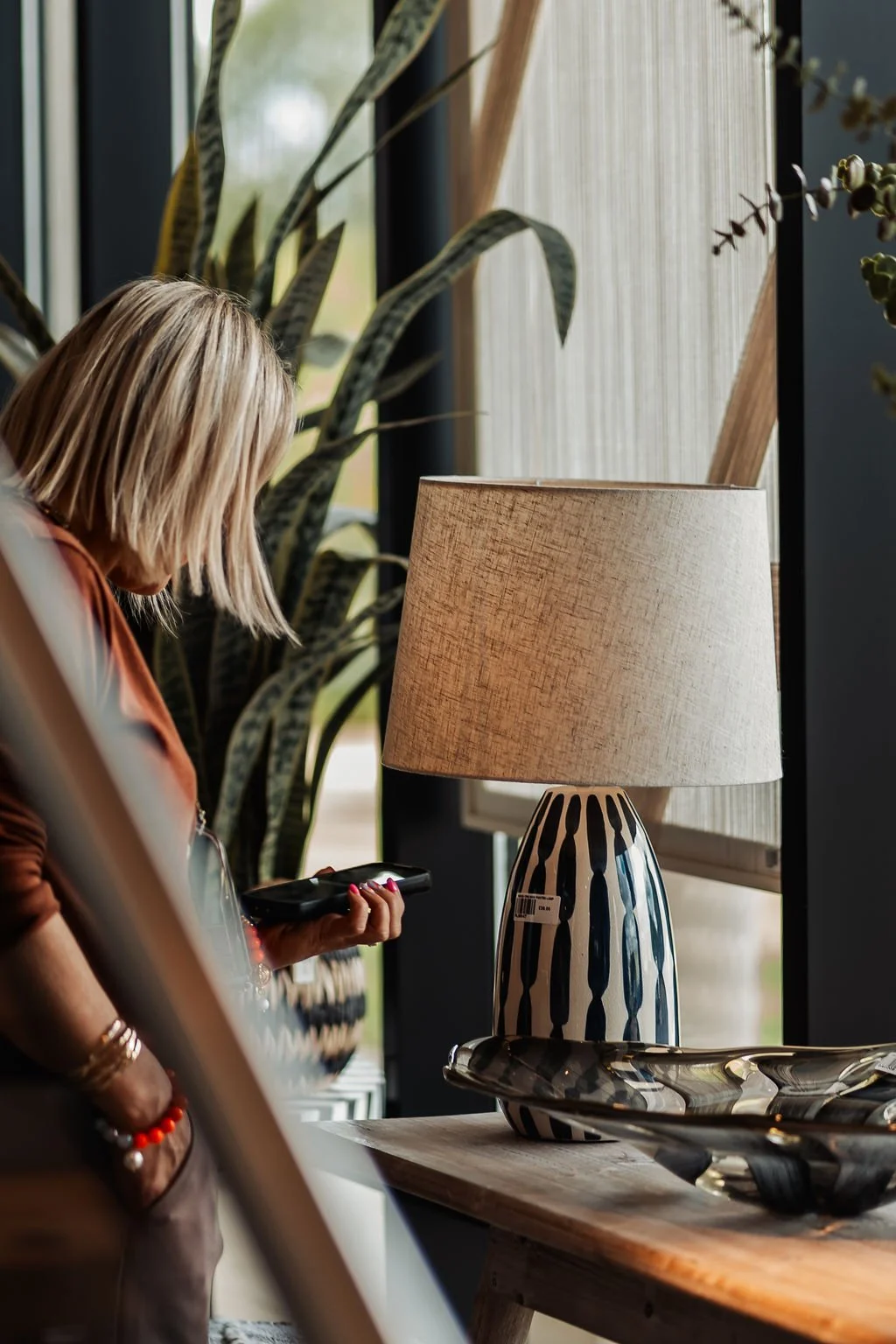 A woman with blonde hair looking at her phone in a decor store with a large striped table lamp, plants, and a wooden table.