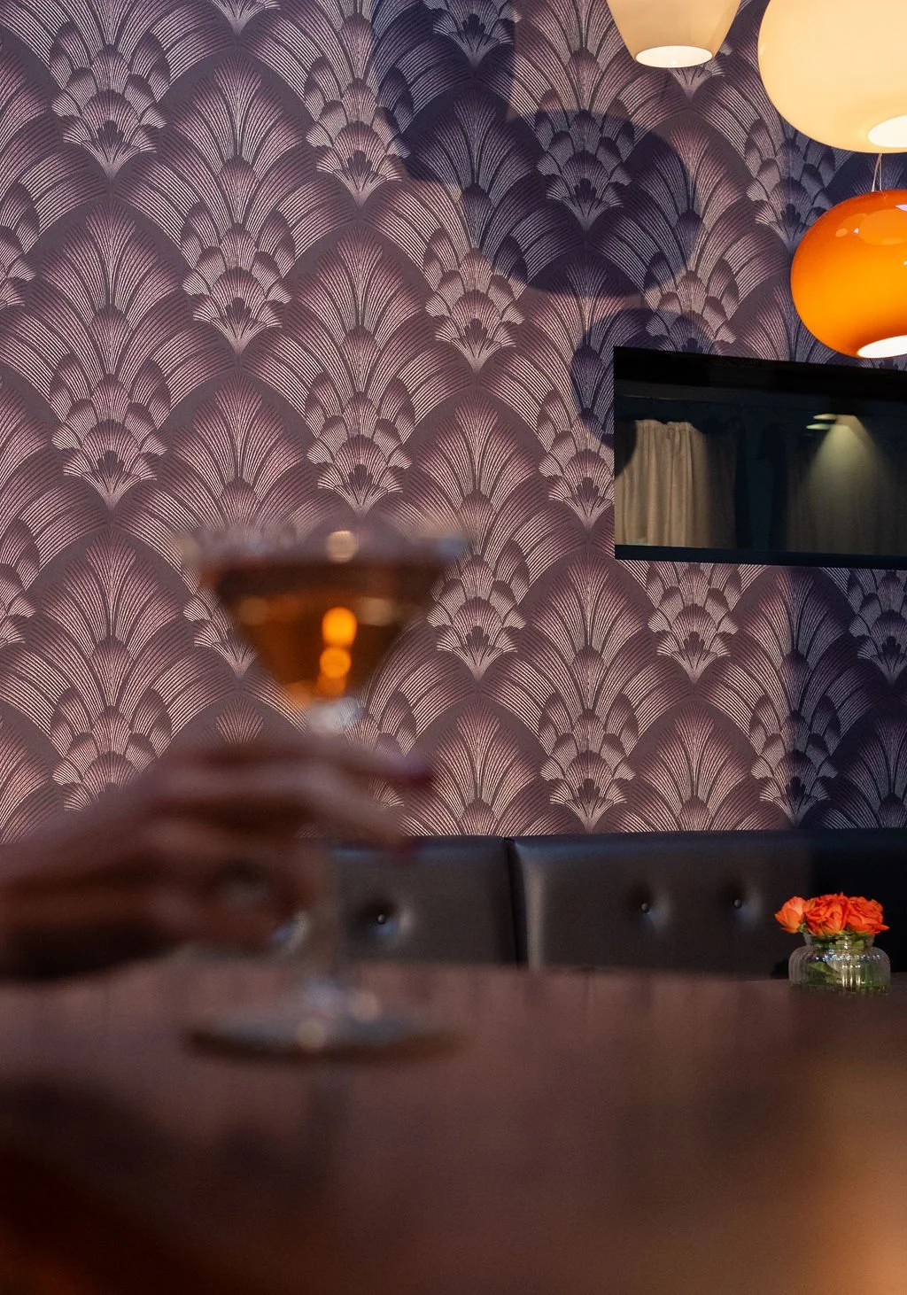 A hand holding a cocktail glass with an orange drink in front of a purple patterned wall in a dimly lit lounge or bar, with a vase of orange flowers on the table and hanging lamps.