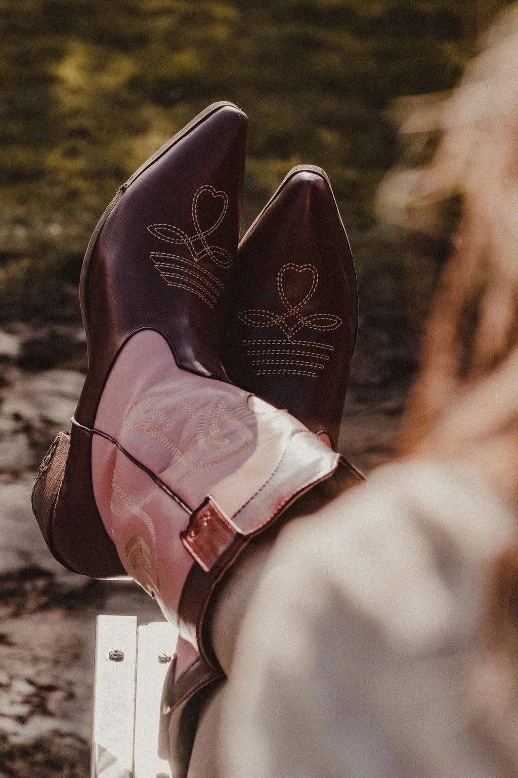 A person wearing pink and burgundy cowboy boots with stitched heart designs, sitting on the bed of a truck.