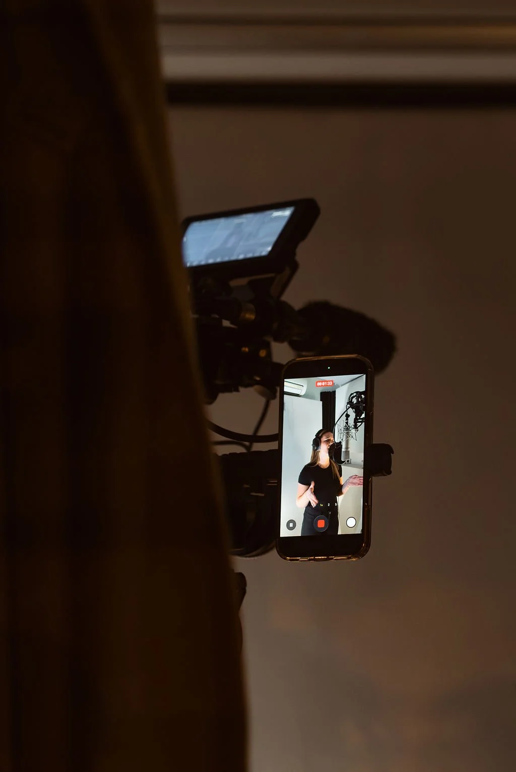 A woman recording a video in a studio is seen on a smartphone screen, with a microphone overhead and a pop-up window showing the live recording.