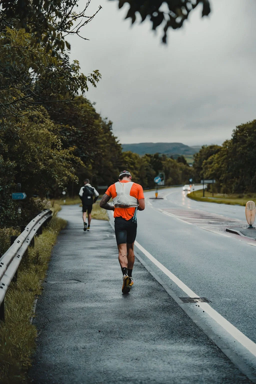 A person running on a wet sidewalk next to a road under cloudy skies, with trees on both sides and another person jogging ahead.