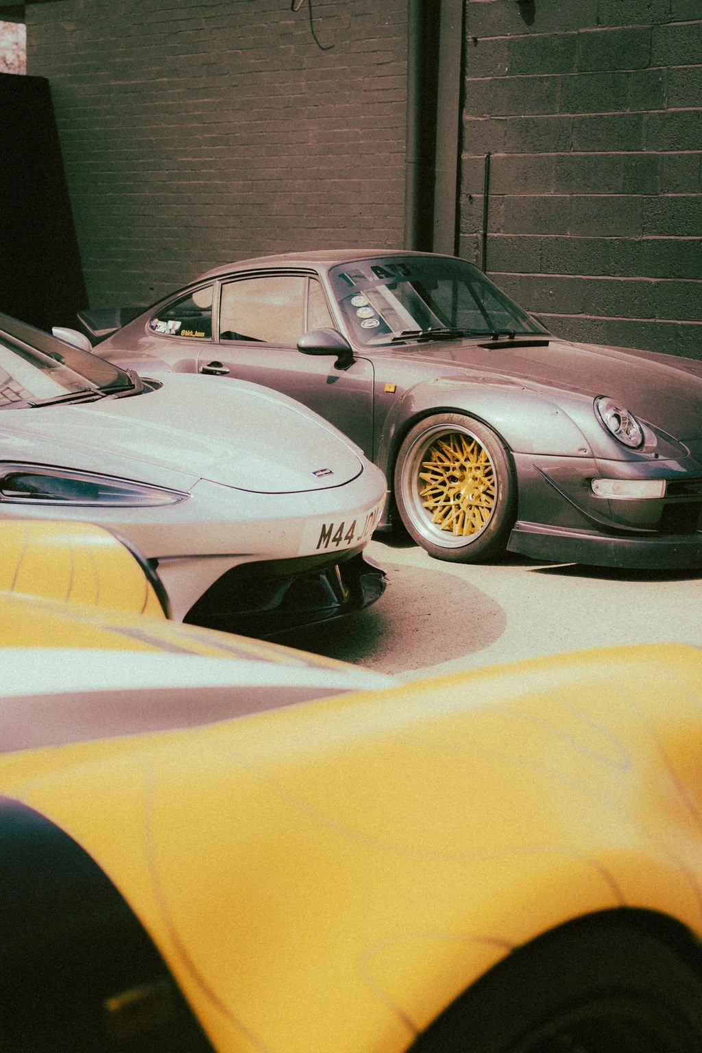 Several cars parked indoors, including a silver Porsche with yellow rims, a white sports car with partial visible front, and a yellow vehicle in the foreground, all against a dark brick wall.