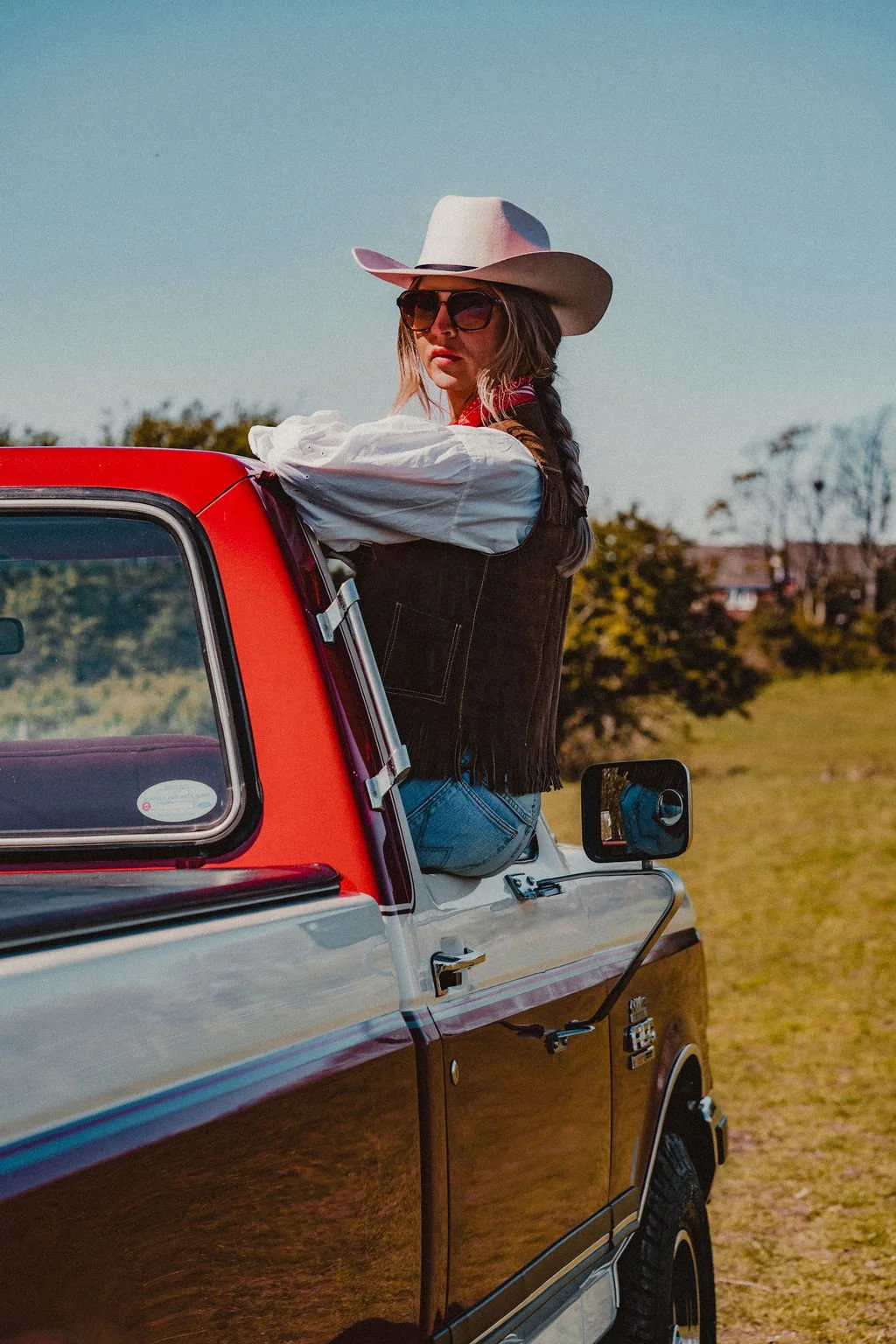 Woman wearing cowboy hat and sunglasses sitting on the door of a vintage pickup truck in a rural outdoor setting.