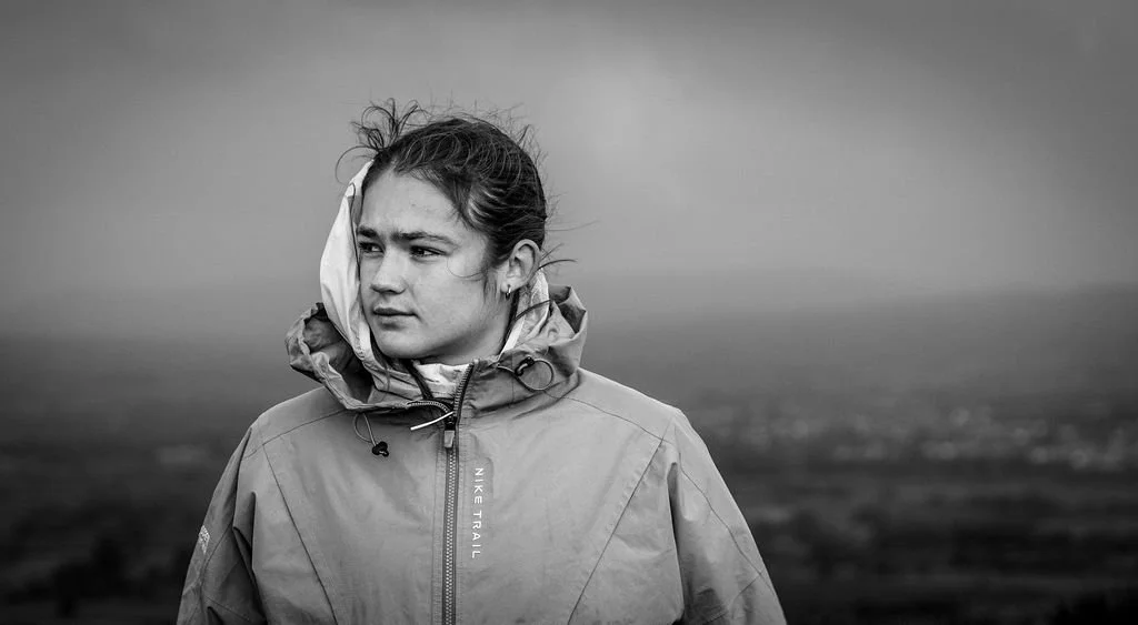A young woman in a Nike Trail jacket standing outdoors on a cloudy day, looking to the side with a contemplative expression.