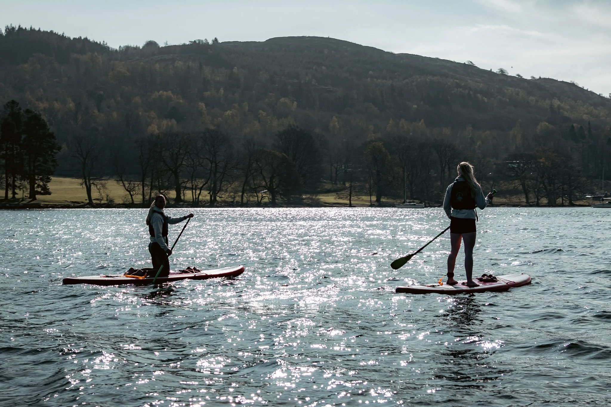 Two people stand on paddleboards on a lake, with a landscape of trees and hills in the background, during a sunny day.