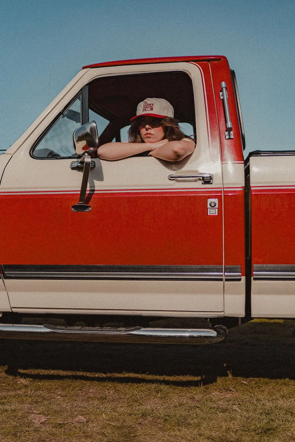 A young woman with brown hair resting her arms on an open window of a red and white pickup truck, wearing a gray baseball cap with red lettering.