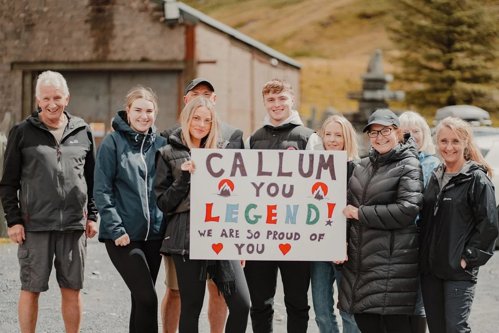 Group of eight people holding a sign that reads "Callum you’re a legend! We are so proud of you," outdoors during daytime with trees and a building in the background.