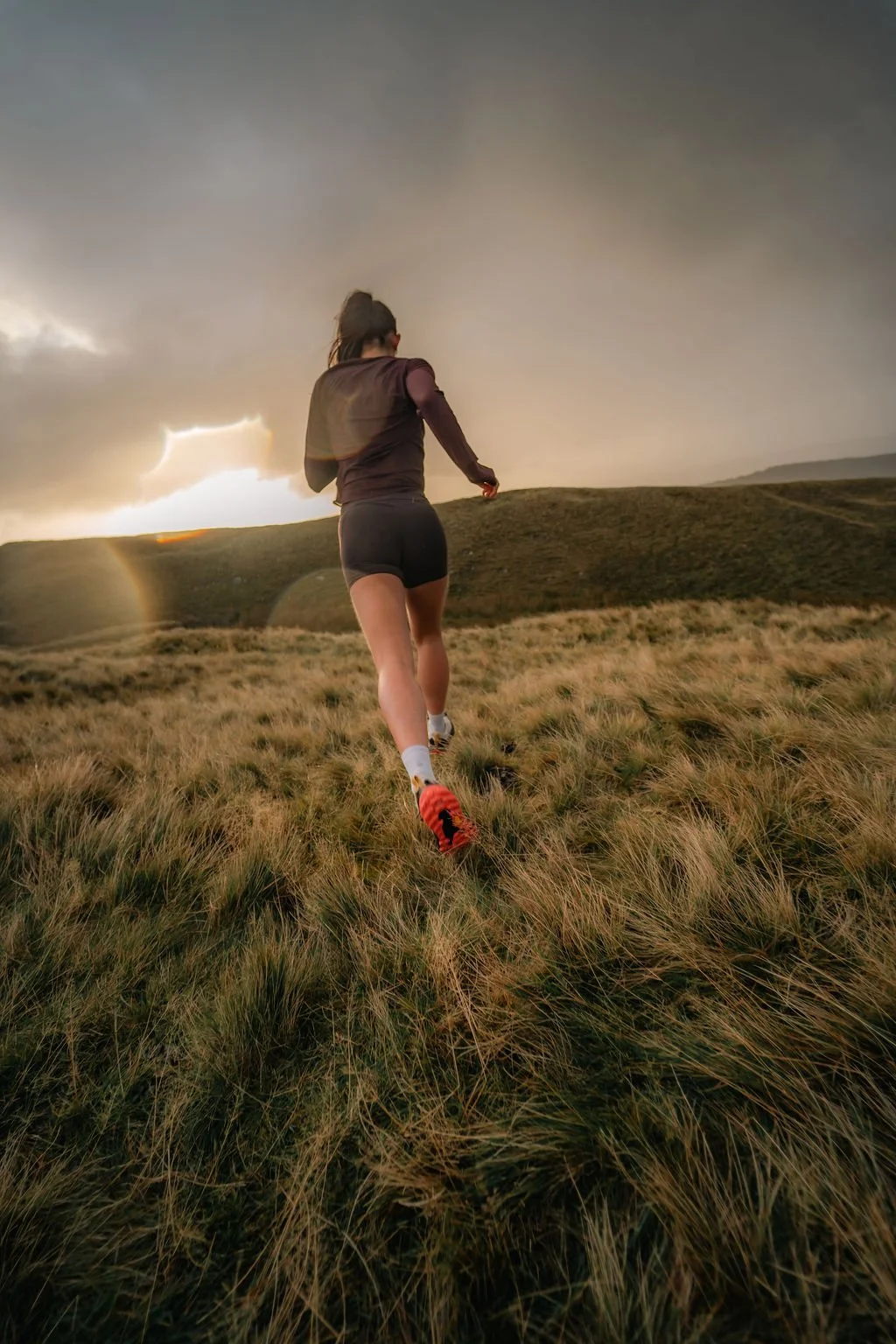 A woman running through grassy landscape during sunset or sunrise, under a partly cloudy sky.
