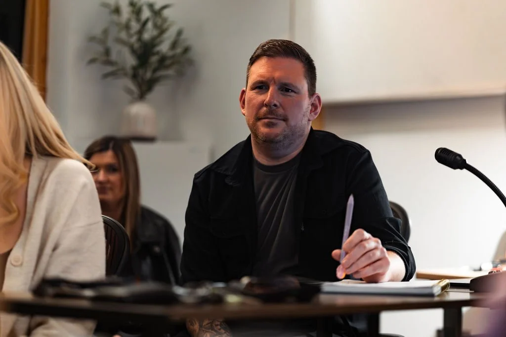 A man sitting at a table with a pen in his hand, looking contemplative, in a meeting or conference room.